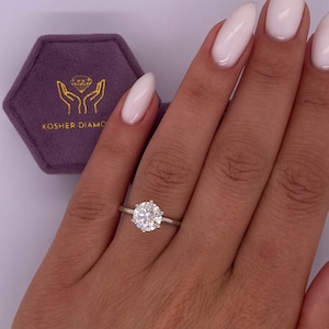 May include: A close-up of a hand wearing a gold ring with a large, round, clear diamond. The ring is set in a simple, classic style. The hand is resting on a purple velvet box with the words "Kosher Diamo" printed on it.