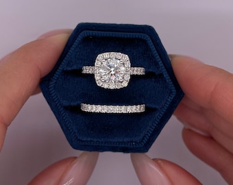 May include: A diamond engagement ring set in a dark blue velvet ring box. The engagement ring features a large, round center stone surrounded by a halo of smaller diamonds. A matching diamond band sits below. The ring box is held by a person.