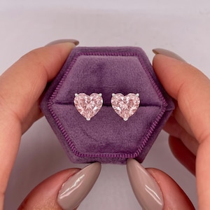May include: A pair of heart-shaped stud earrings featuring light pink faceted stones set in silver-toned metal prongs. The earrings are displayed inside a hexagonal purple velvet jewelry box with a single slot for each earring.
