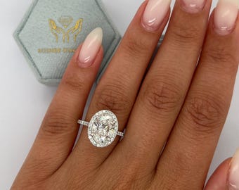 Puede incluir: Un anillo de plata con un gran diamante de forma ovalada rodeado por un halo de diamantes más pequeños. El anillo está en un dedo.