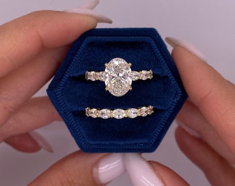 Könnte beinhalten: Ein Paar Verlobungsringe in einer blauen Samt-Ringbox. Der Hauptring hat einen großen ovalen Diamanten mit kleineren Diamanten auf der Ringschiene. Der zweite Ring ist eine Band aus kleineren ovalen Diamanten.