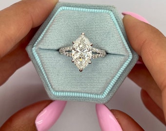 Puede incluir: Un anillo de compromiso de diamante de forma de pera con un halo doble. El anillo está engastado en una banda de oro blanco y se muestra en una caja de anillo de terciopelo azul claro.