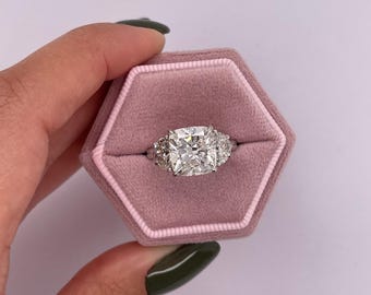 May include: A large, square-shaped diamond engagement ring with smaller diamonds on the band. The ring is in a pink velvet ring box.