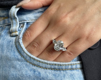 May include: A close-up of a hand wearing a silver ring with a large oval-shaped diamond surrounded by smaller diamonds.