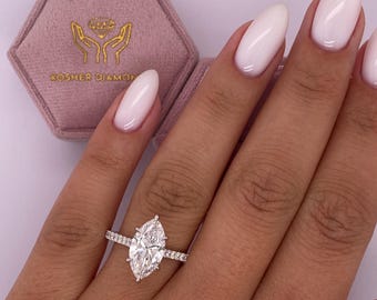 Puede incluir: Primer plano de una mano con un anillo de compromiso de diamante de talla marquesa con una banda de diamantes pavé. El anillo está engastado en oro blanco. El anillo está en una caja de anillo de terciopelo rosa con las palabras "Kosher Diamond" impresas en la caja.