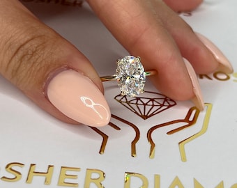 Può includere: Primo piano di una mano che indossa un anello d'oro con un grande diamante di forma ovale. L'anello è incastonato in una semplice fascia e il diamante brilla.