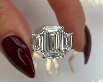 May include: A three-stone emerald cut diamond engagement ring with a large center stone and two smaller stones on either side. The ring is set in a white gold band.