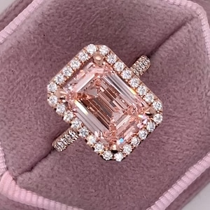 Könnte beinhalten: Ein Ring aus Roségold mit einem großen, rechteckigen, pfirsichfarbenen Mittelstein im Smaragdschliff, der von einem Kranz aus kleinen, runden, klaren weißen Steinen umgeben ist. Die Ringschiene ist ebenfalls mit kleinen, runden, weißen Steinen besetzt.