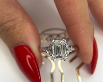 May include: A silver engagement ring featuring an emerald cut diamond centre stone and a halo of smaller diamonds. The ring is on a finger with red nail polish.