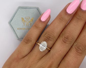 May include: An oval-cut solitaire diamond ring set on a thin gold band. The ring is positioned in front of a light blue hexagonal velvet jewelry box featuring a gold logo of two hands holding a diamond and the text POSHER DIAMOND.