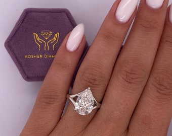 Puede incluir: Un anillo de compromiso de diamante en forma de pera con una montura de doble banda. El anillo está en el dedo de una persona y el diamante brilla.
