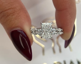 May include: A close-up of a diamond engagement ring with a large, rectangular-shaped center stone and a delicate, pave-set band. The ring is set in white gold and features a halo of smaller diamonds around the center stone.