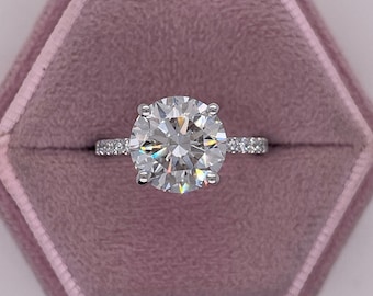 May include: A sparkling diamond engagement ring with a round brilliant cut center stone and a pave diamond band. The ring is set in a white gold setting and is displayed in a pink velvet ring box.