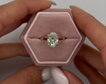 Könnte beinhalten: Ein goldener Ring mit einem ovalen Diamanten, der in einer Krappenfassung sitzt. Der Ring befindet sich in einer rosafarbenen Samt-Ringbox.