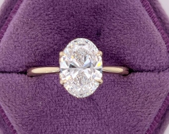 Könnte beinhalten: Ein goldener Bandring mit einem großen, oval geschliffenen Diamanten. Der Diamant ist in einer klassischen Krappenfassung gefasst und funkelt hell. Der Ring wird in einer lila Samt-Ringbox präsentiert.