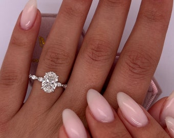 May include: A close-up of a hand wearing a silver ring with an oval-shaped diamond and smaller diamonds on the band. The hand has a light pink manicure with almond-shaped nails.
