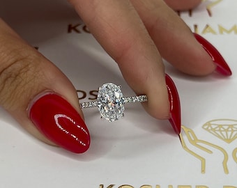 May include: A silver solitaire engagement ring with a large oval-shaped diamond set in a pave band. The ring is on a finger with red nail polish.