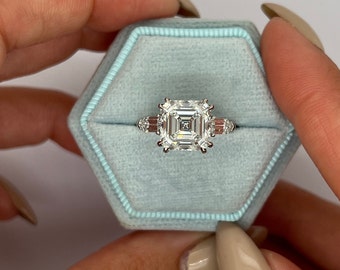May include: A square-shaped diamond engagement ring with a white gold band and smaller diamonds on the sides. The ring is in a light blue velvet ring box.