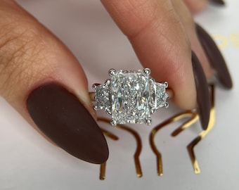 May include: A silver ring featuring a large, square-shaped diamond in the centre. The diamond is surrounded by smaller diamonds on either side. The ring is on a white surface with the words "Sher Diam" in gold lettering.