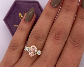 May include: A gold ring featuring a large, light pink marquise-cut center stone, flanked by two smaller, round, clear stones. The ring is set on a hand with brown nail polish, next to a purple velvet ring box.