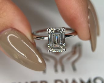 Puede incluir: Un anillo de plata con un diamante de talla esmeralda. El diamante es transparente y tiene mucho brillo. El anillo está en un dedo con esmalte de uñas marrón claro.