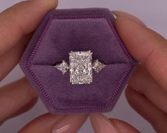 May include: A radiant cut diamond engagement ring with three smaller diamond accents. The ring is set in a white metal band and is displayed in a purple velvet ring box.