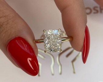 Op de afbeelding: Een gouden ring met een grote, kussen geslepen diamant. De diamant is gezet in een kraalbezetting en heeft veel glans. De ring zit aan een vinger met rode nagellak.