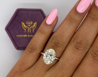 May include: A close-up of a hand wearing a ring with a large oval-shaped diamond. The ring is set in a simple band and the diamond is sparkling. The ring is on a finger with pink nail polish. The background is a white surface with a purple velvet box with the text "Kosher Diam" on it.