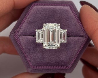 May include: A three-stone emerald cut diamond engagement ring with a large center stone and two smaller side stones. The ring is set in a white gold band and is displayed in a purple velvet ring box.