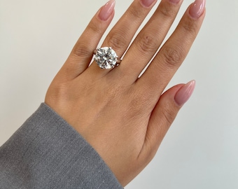 May include: A large, round-cut diamond ring with a silver band. The ring is set on a hand with manicured nails, and the sleeve of a grey blazer is visible in the frame.