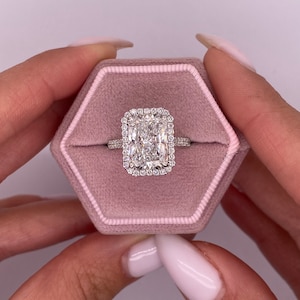 Könnte beinhalten: Ein quadratischer Diamantring mit Halo-Fassung. Der Ring ist in einer weißen Metallfassung gefasst und wird in einer rosafarbenen Samt-Ringbox präsentiert.