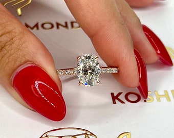 Puede incluir: Anillo de compromiso de oro rosa con un diamante central de talla ovalada. La banda está adornada con diamantes más pequeños engastados en pavé. Sostenido por una mano con esmalte de uñas rojo, sobre un fondo blanco con detalles dorados en forma de diamante.