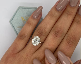 May include: A silver engagement ring with a large oval-cut diamond. The ring is set on a hand with long, almond-shaped nails painted in a neutral color. A light blue hexagonal ring box is in the background.