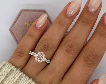 May include: A silver ring with a large oval-cut pink gemstone, flanked by smaller white gemstones. The ring is on a person's finger, with a light pink velvet ring box in the background.