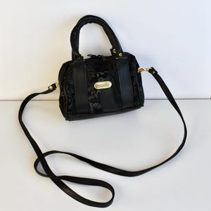 Könnte beinhalten: Schwarze Ledertasche mit einem goldfarbenen Logo mit der Aufschrift "Stefano". Die Tasche hat einen oberen Henkel und einen langen verstellbaren Riemen.
