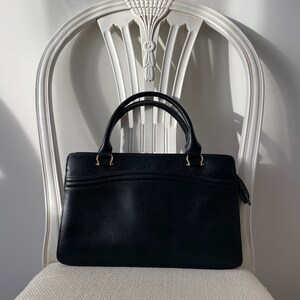 May include: A black leather handbag with gold hardware and a top handle. The bag is sitting on a white chair.