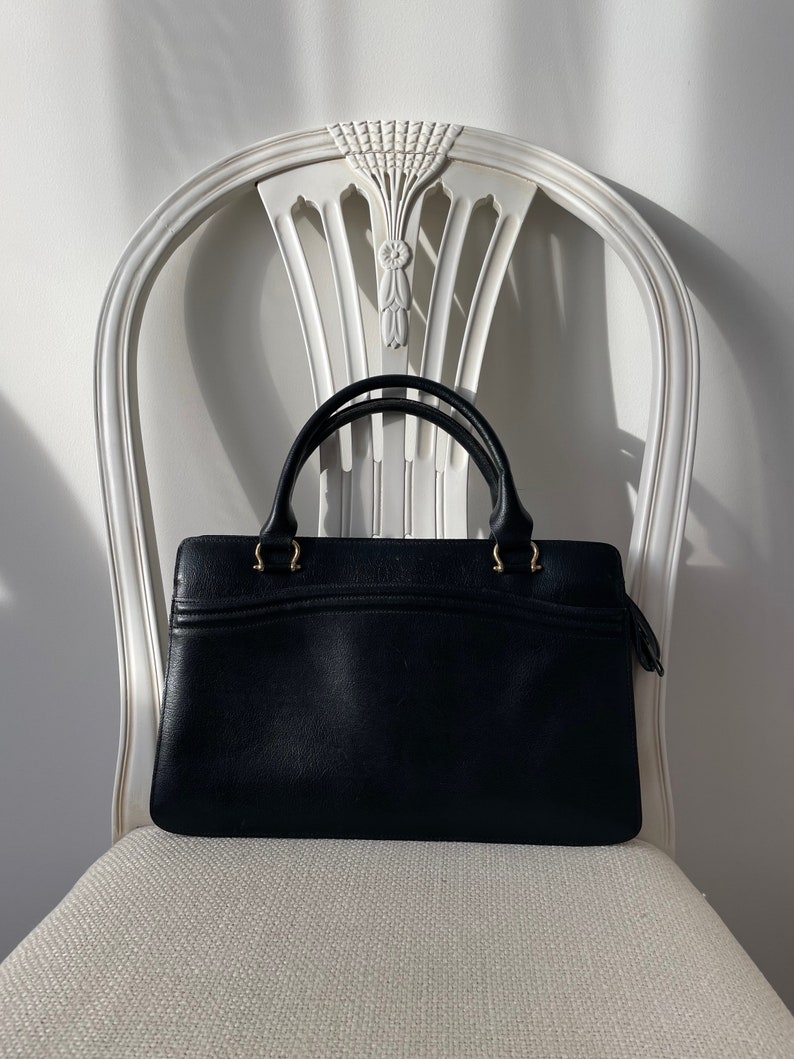 May include: A black leather handbag with gold hardware and a top handle. The bag is sitting on a white chair.