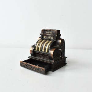 May include: A vintage National brand metal pencil sharpener shaped like a cash register. The sharpener has a drawer that pulls out and a display that reads "Contado Preços - Cta 00.00".