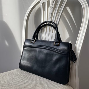 May include: A black leather handbag with gold hardware and a zipper closure. The bag is sitting on a white chair.