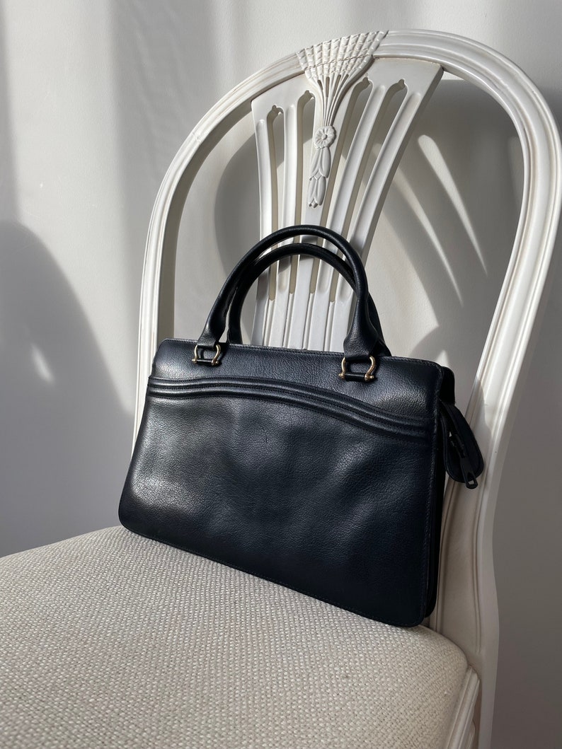 May include: A black leather handbag with gold hardware and a zipper closure. The bag is sitting on a white chair.