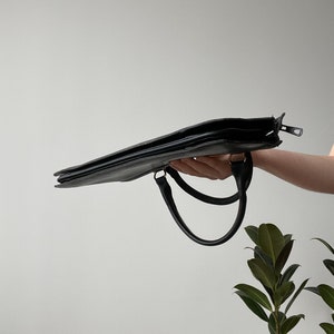 May include: A black leather briefcase with two handles. The briefcase is held in a hand and the zipper is open.