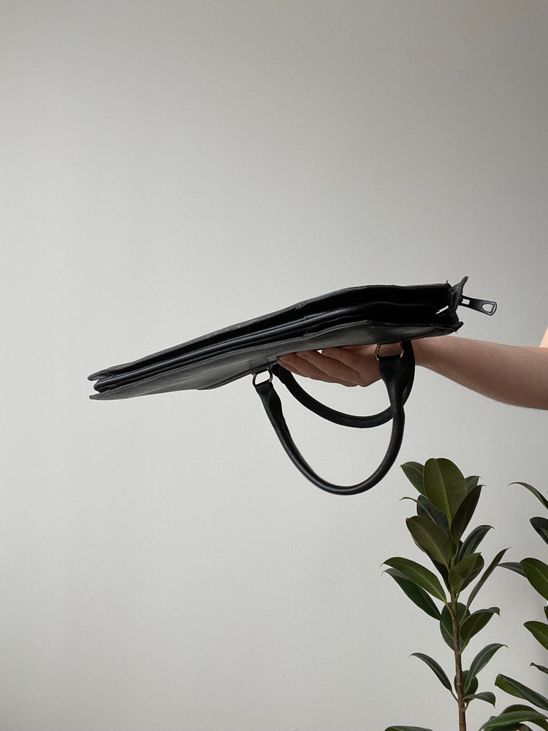 May include: A black leather briefcase with two handles. The briefcase is held in a hand and the zipper is open.