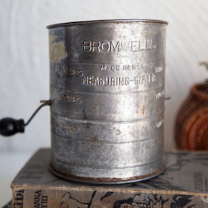 Pode incluir: Peneira de medição de metal vintage com o texto "BROMWELL'S MADE IN USA MEASURING-SIFTER". Possui uma alça preta e marcações de medição para xícaras. A peneira está em cima de uma pilha de livros antigos. Feito nos EUA.