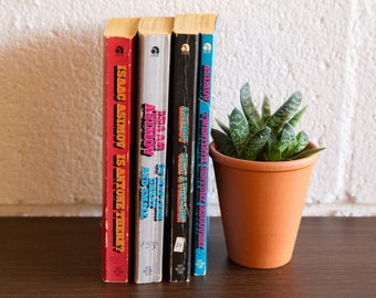 May include: Four vintage paperback books by Isaac Asimov, with titles including "Is Anyone There?" and "Get a Thrill". The books are in various colors, including red, silver, black, and blue. A small potted succulent plant sits to the right.