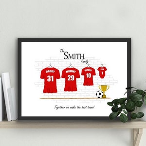 May include: A framed print featuring a family's soccer jerseys, each with a name and number. The red jerseys hang against a white brick wall, with a soccer ball and trophy. The family name is at the top, and the phrase "Together we make the best team!" is at the bottom.