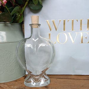 May include: A heart-shaped glass bottle with a cork stopper. The bottle is on a wooden surface, with a light blue background that reads "WITH LOVE" in gold lettering. A green vase with flowers is in the background. The bottle is likely for decorative use.