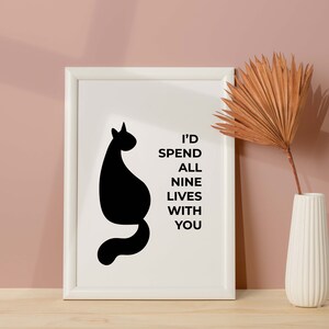May include: A white framed print with a black silhouette of a cat and the text "I'd spend all nine lives with you". The print is on a wooden surface with a dried palm leaf in a white vase.
