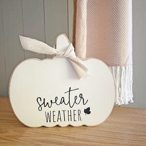 May include: A white pumpkin-shaped wooden sign with a light beige fabric ribbon tied around the stem. The sign features the black text "sweater WEATHER" and a small black leaf icon printed on the front surface, showcasing a smooth finish.