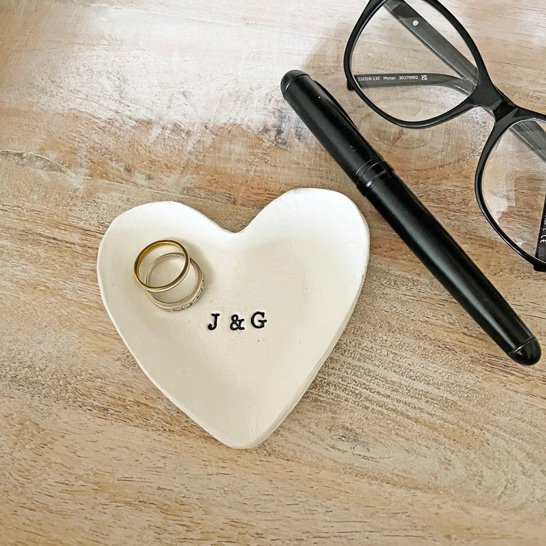 May include: A white heart-shaped ring dish with the initials "J & G" in black lettering. Two rings, one gold and one silver, rest inside the dish. A black pen and black-framed glasses are also visible.