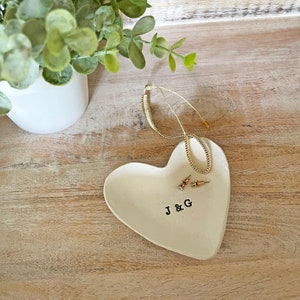 May include: A heart-shaped, off-white ceramic jewelry dish with the initials "J & G" stamped in black. A gold necklace and a pair of stud earrings rest on the dish. A small white pot with greenery is in the background.